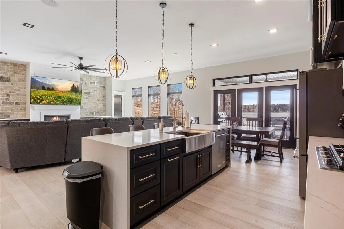 The image captures a stunning open-concept kitchen and living area, centered around a sleek kitchen island with modern pendant lighting. Expansive windows frame serene lake views, and a cozy seating area invites guests to relax, with warm lighting enhancing the inviting ambiance.