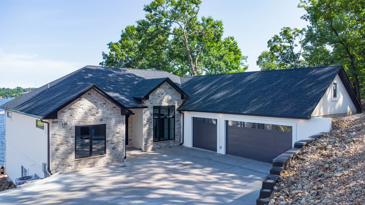A welcoming exterior angle of Grand Gaze reveals its inviting stone façade, expansive windows, and smooth driveway. The home’s proximity to the water and the potential for outdoor amenities suggest an ideal environment for both focused work sessions and leisure activities, such as morning coffee on the patio or evening bonfires.