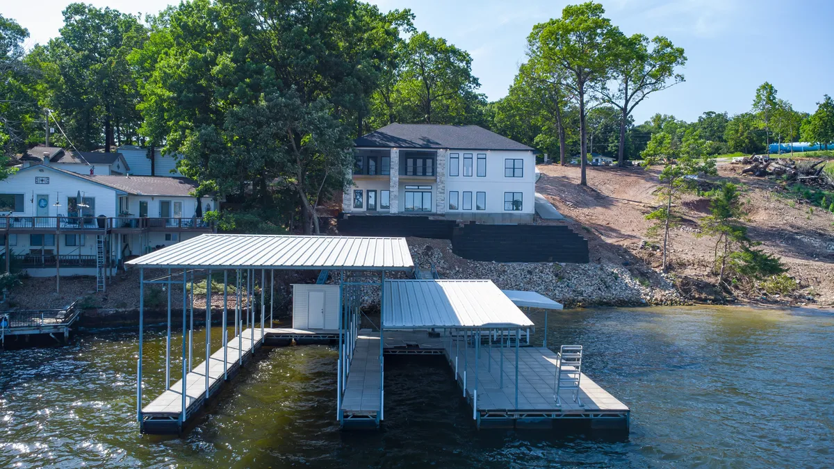This image presents a stunning lakefront scene featuring a boathouse, the modern architectural style of the home, and inviting outdoor areas. The scene exudes a relaxed, vacation-like atmosphere with space for sunbathing, gathering, and engaging in water activities like boating and fishing. The composition centers on the connections to the water and the property's private setting.