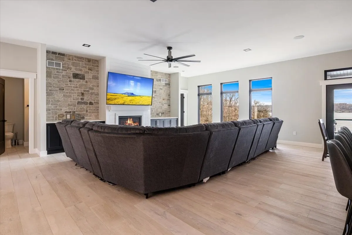 A spacious, inviting living room featuring a large sectional sofa, a modern fireplace, a wall-mounted television, and expansive windows with lake views. The setting is perfect for group relaxation, movie nights, or sharing stories by the fire.
