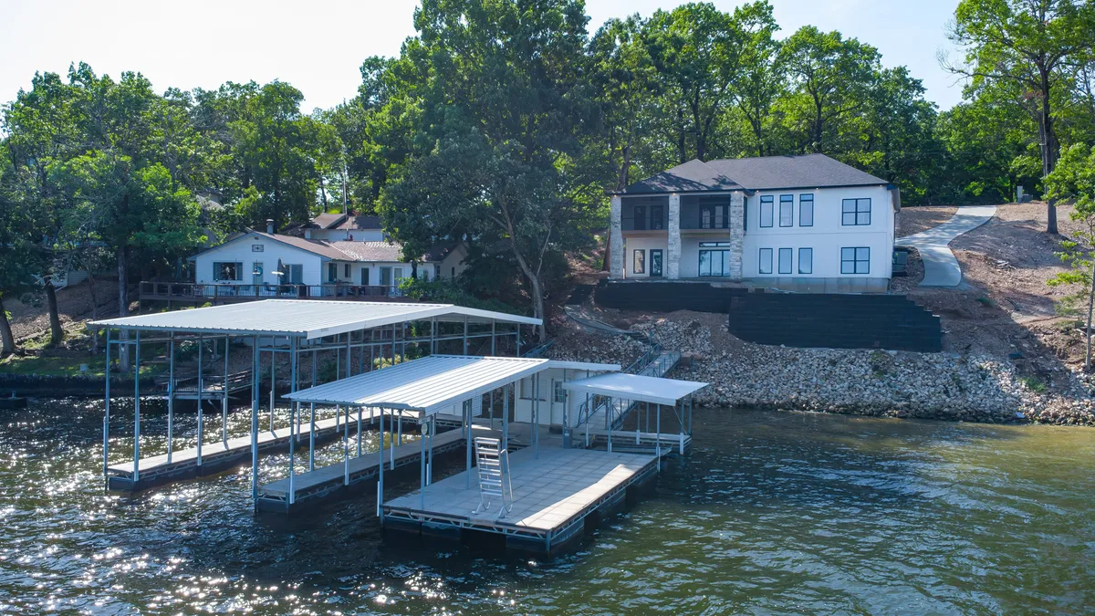A tranquil waterfront scene featuring a modern house with a prominent dock jutting over the lake. The dock is large enough for boat and PWC slips, surrounded by lush trees and calm water, evoking a sense of privacy and lakeside leisure.