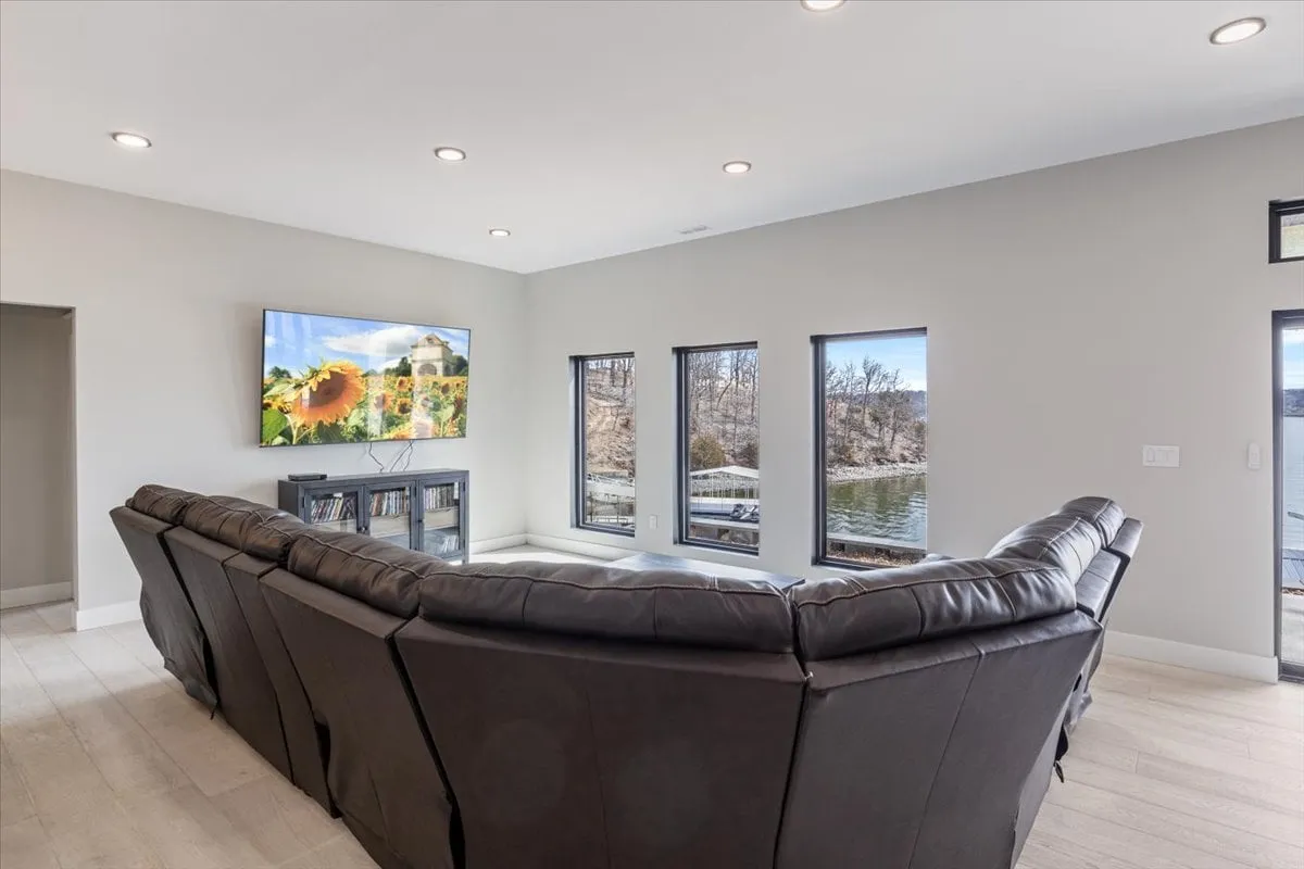 A cozy lounge setting with a large sectional sofa facing a TV displaying vibrant imagery and expansive windows overlooking a serene lakeside scene. The mood is relaxed and ideal for gatherings or movie marathons.